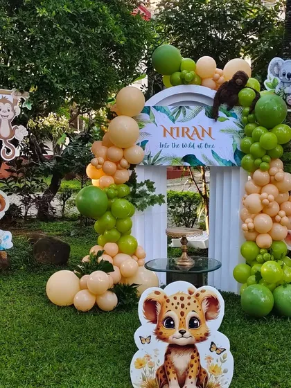 We transformed this garden into a wild jungle adventure for a first birthday. The setup includes an organic balloon arch in earthy tones, a custom 'Into the Wild at One' backdrop, and adorable cutouts of jungle animals like lions, monkeys, and giraffes.