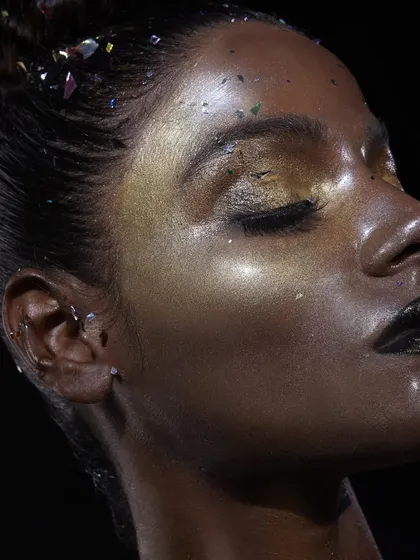 This profile view showcases the incredible shine and texture of the glass skin technique. The light catches the gold highlighter on the cheekbones and the glitter on the eyelids, creating a futuristic and ethereal party look.