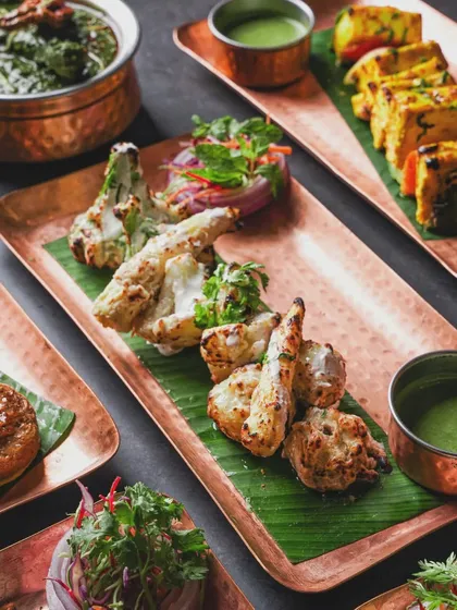 A spread of North Indian appetizers, including paneer tikka and malai cauliflower, served on copper platters. The dark background makes the colors of the food and green chutney pop.