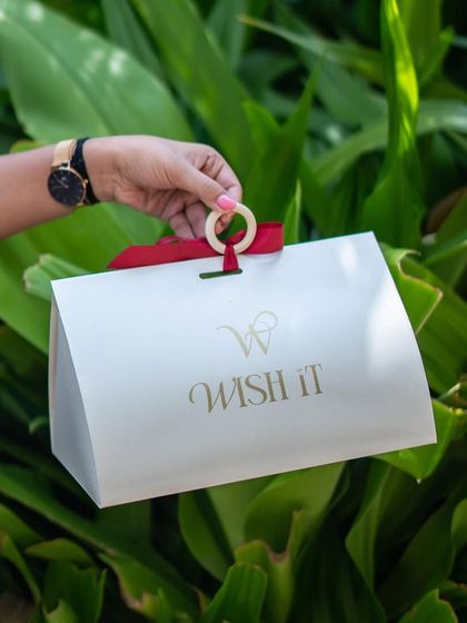 Holding our "Wishlet Kit" against a backdrop of lush greenery. The clean, white design with a pop of red ribbon makes it a stylish and modern gift bag.