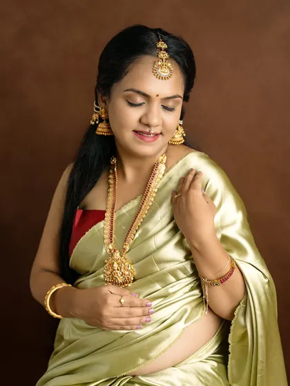 A close-up portrait of a beautiful mom-to-be adorned in traditional gold jewelry and a silk saree, looking down with a gentle smile.