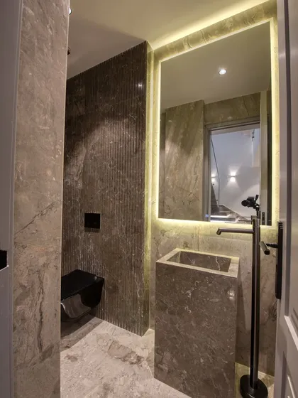 This powder room features a monolithic stone pedestal sink and a floor-mounted faucet. The back-lit mirror and fluted stone accent wall create a sophisticated, moody atmosphere.