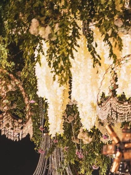 An intricate ceiling installation featuring a dense canopy of greenery, white wisteria, and crystal hangings. The mix of textures and materials creates a luxurious and enchanting overhead view.