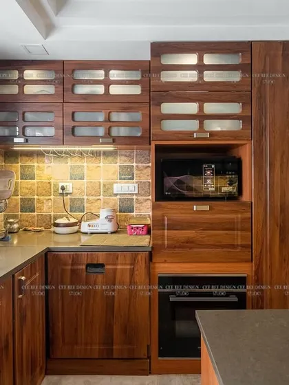 A luxurious kitchen with rich teakwood veneer cabinets, frosted glass shutters, and an integrated appliance unit.