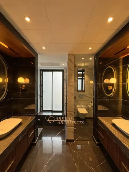 A bold and dramatic bathroom with black marble walls, dual vanities, and warm wood accents. The backlit oval mirrors add a soft, glamorous glow.