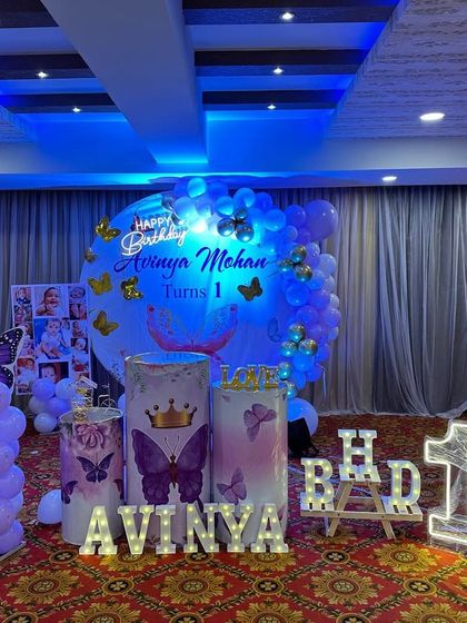 A butterfly theme with a royal touch, using purple and white balloons and gold crown details. The setup includes themed plinths, balloon columns, and marquee letters for the name and age.