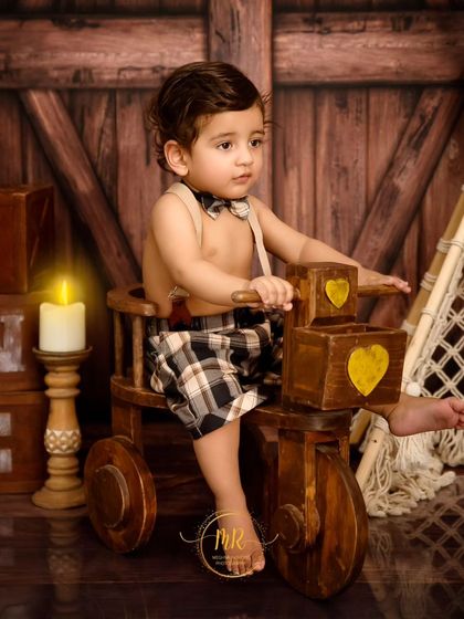 A one-year-old boy in plaid shorts and a bow tie, sitting on a wooden tricycle prop in a rustic barn-themed birthday setup.