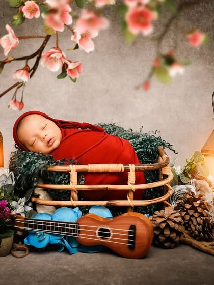 A sleeping newborn in a red wrap, surrounded by cherry blossoms, a ukulele, and warm lanterns. This setup combines rustic and musical elements for a unique portrait.