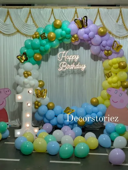 A vibrant Peppa Pig first birthday party, complete with a full balloon ring in pastel rainbow colors. The setup is brought to life with a 'Happy Birthday' neon sign, a light-up marquee number '1', and character cutouts of Peppa and George.