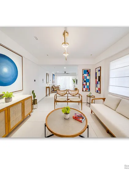 A contemporary living room with a clean, all-white aesthetic, punctuated by bold, colorful artwork. The natural wood and rattan furniture add warmth and texture, creating a space that feels both modern and inviting.