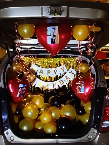 A unique car trunk birthday surprise, perfect for creating a memorable moment. This setup uses gold, black, and red balloons, a "Happy Birthday" banner, and personalized photos attached to heart-shaped foil balloons.