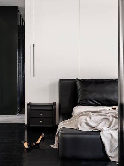 The master bedroom in the Ebony & Ivory project is a statement in moody minimalism. A low-profile black leather bed is set against a stark white wall, with a sleek black nightstand and a single pendant light.