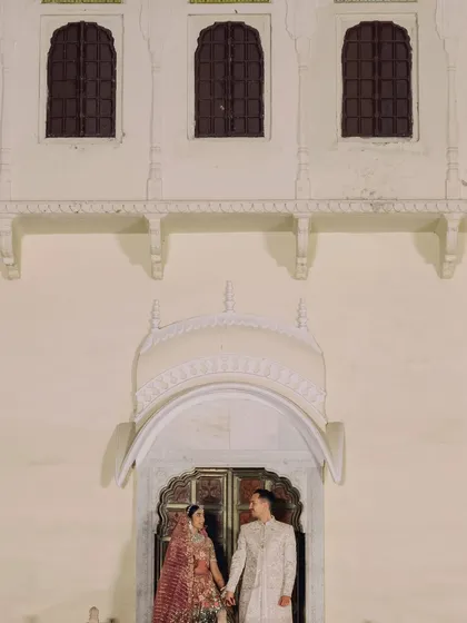 The couple shares a private moment, holding hands in a doorway against the grand facade of the palace, showcasing the scale of the venue.