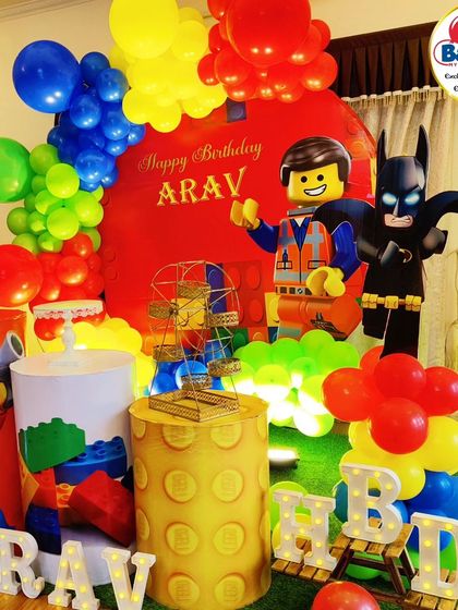 A primary-colored LEGO-themed 10th birthday party. The setup includes LEGO Batman and Emmet cutouts, LEGO-brick plinths, and a 'ARAV' marquee.