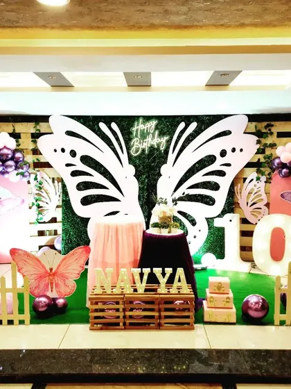 This butterfly-themed stage for Navya's 10th birthday features a lush greenery wall as the base. I added dramatic, oversized white butterfly wings as the central photo-op element, framed by balloon columns in shades of purple and pink for a vibrant, garden-like feel.