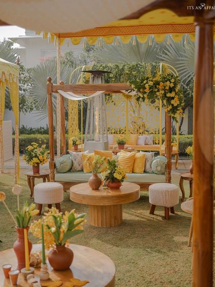 A cozy seating area under a yellow tent for the 'Bageecha' Haldi, featuring wooden furniture, soft green cushions, and pops of yellow.