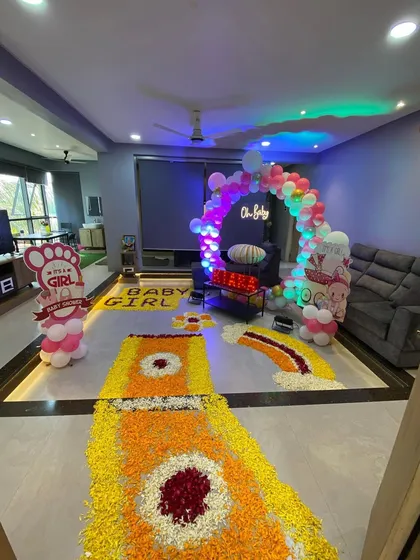 A full view of a living room decorated for a baby's homecoming, with a flower rangoli path, a balloon arch, and a neon 'Oh Baby' sign.