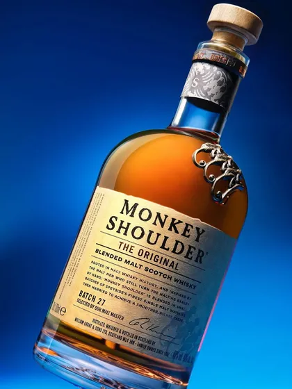 A classic bottle shot of Monkey Shoulder whiskey, using a tilted angle to create a more dynamic composition. The clean blue background and sharp lighting make the bottle and its label stand out clearly.