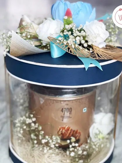 A wedding snacks hamper featuring a tin of 4700BC popcorn, presented in a stylish cylindrical box with a blue and white floral arrangement on top.
