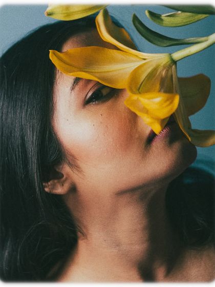 Using props like flowers can add a layer of poetry to a beauty shot. Here, the yellow lily partially obscures the model's face, creating a sense of mystery and artistry. The cool blue background contrasts beautifully with the warm tones of the flower and skin.