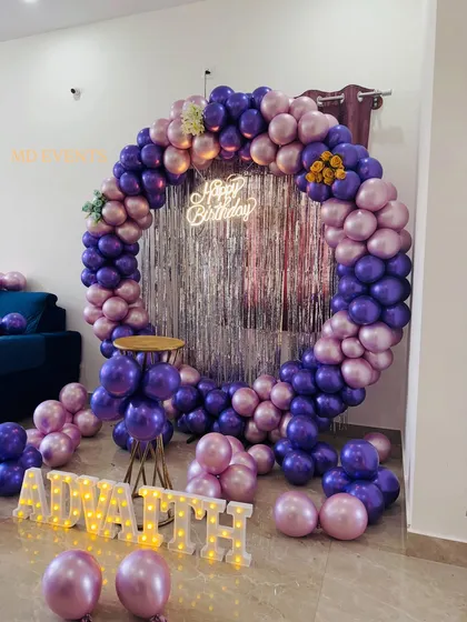 I love working with bold color palettes. This birthday decoration uses a circular frame wrapped in metallic purple and pink balloons, set against a silver fringe curtain for extra sparkle. The setup is completed with marquee letters for a personalized touch.