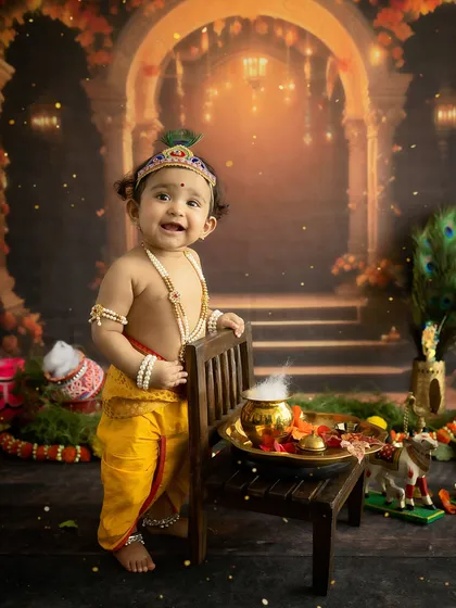 A full-length portrait of a baby dressed as Little Krishna for Janmashtami, standing by a chair with a pot of "butter."