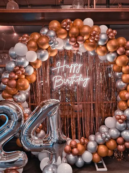 A chic 24th birthday setup with a circular balloon arch in copper, silver, and white. The decoration is set against a fringe backdrop with a neon sign.