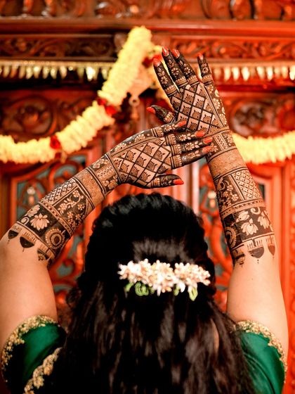 A beautiful bride in a green saree, her hands covered in intricate geometric and floral mehendi. The back-of-hand design is just as stunning as the front.
