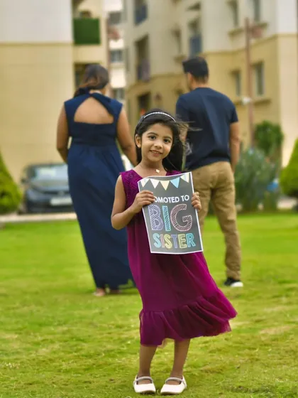 An exciting family announcement, with a young girl proudly holding a "Promoted to Big Sister" sign while her parents stand in the background, beautifully capturing the anticipation of a new arrival.