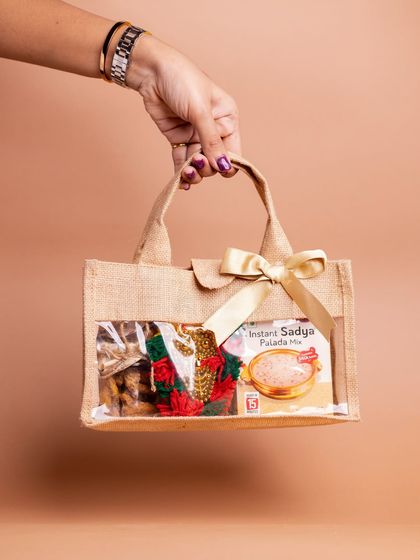 A hand holding the 'Kaithola' jute bag hamper, showcasing its compact and charming presentation. The clear window offers a peek at the festive goodies inside.