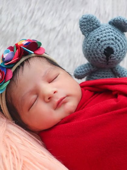 A close-up of a baby girl in a vibrant red swaddle, sleeping peacefully with her teddy bear against a soft pink background.