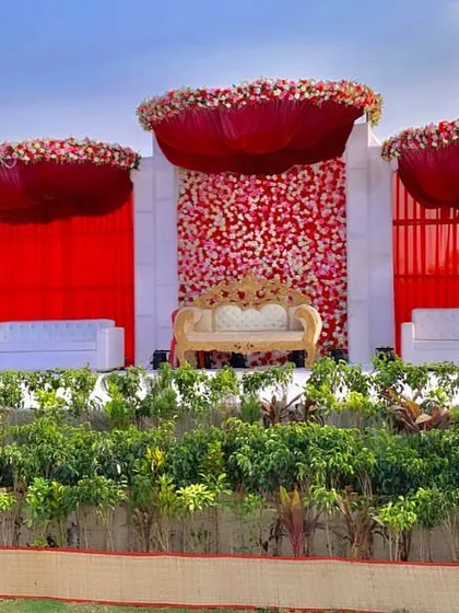 A daytime view of a spectacular outdoor wedding stage. The vibrant red floral arrangements and drapery stand out beautifully against the open sky, creating a powerful and photogenic setting.