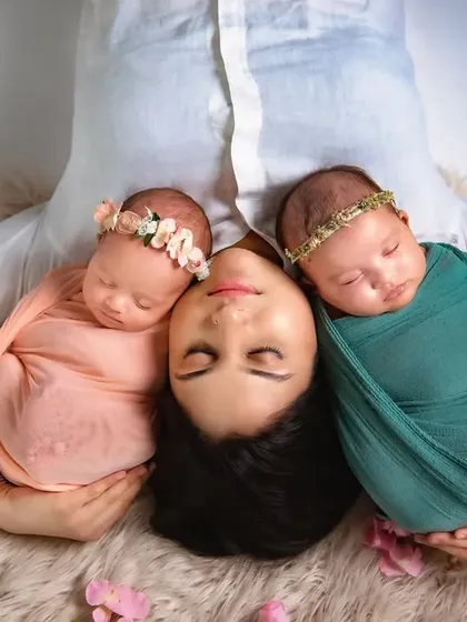 Double the joy. A mother peacefully lies with her twin newborn daughters, each swaddled and resting on her shoulders. I have experience and patience for twin newborn photoshoots.