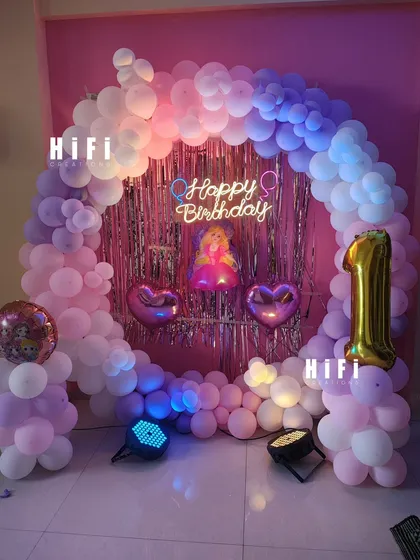 A lovely pastel-themed birthday decoration designed for a cozy home setting. The arch of pink and purple balloons frames a shimmer backdrop, creating a perfect spot for cake cutting.