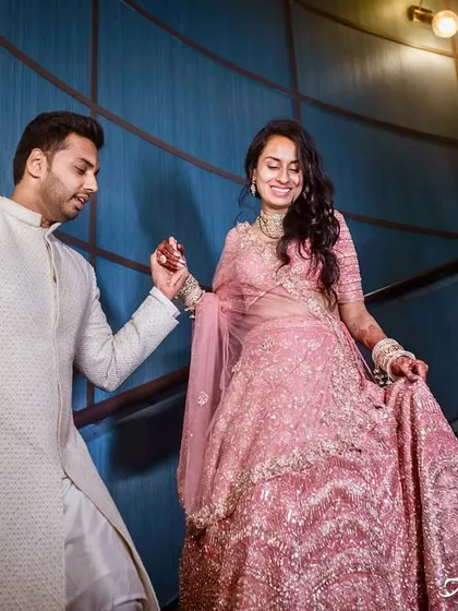 The groom gently leads his bride down a modern, curved staircase. This shot captures movement and elegance, highlighting the beautiful flow of the bride's pink lehenga.