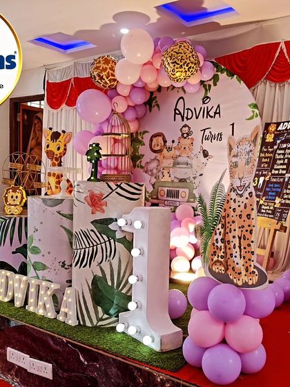 A fun jungle animal party setup for a first birthday. This stage features a variety of animal cutouts including a zebra, leopard, and elephant, with a soft purple and pink balloon garland.
