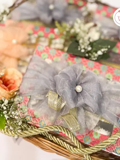 A close-up of a wicker basket hamper, showing intricately wrapped mithai boxes tied with delicate grey organza ribbons.