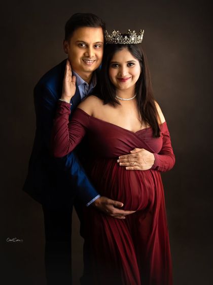 A royal-themed couple's portrait. The mom-to-be wears a crown as her partner stands lovingly behind her, both sharing a smile.