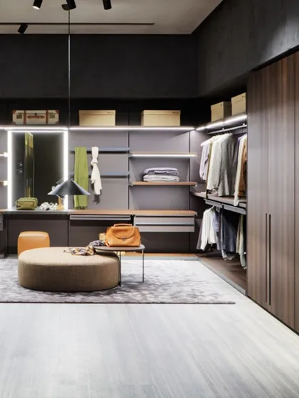 This walk-in closet design uses a dark, masculine color palette with rich wood finishes and an open-plan layout. The integrated vanity and central ottoman create a comfortable and functional space for daily routines.
