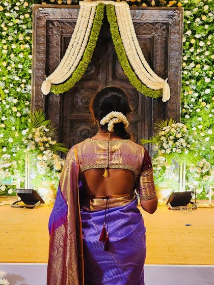 A back view of a client facing a grand, floral-decorated doorway, wearing a purple saree and a brocade blouse with a tie-up back.