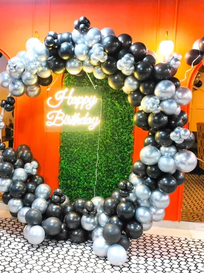 This is a great example of a sophisticated party backdrop for adults. A full ring of black and silver chrome balloons frames a green foliage wall, with a neon "Happy Birthday" sign in the center, creating a perfect spot for selfies and group photos.