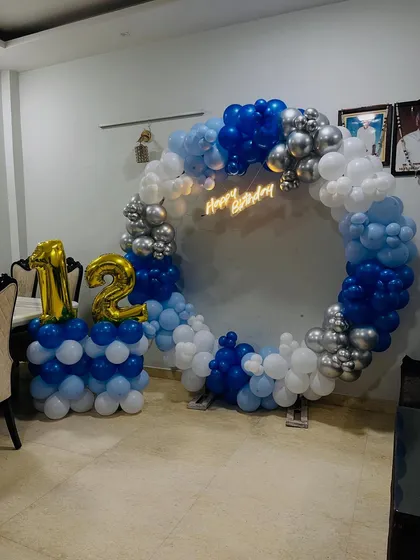 A classic 12th birthday decoration with a full ring of blue, white, and silver chrome balloons, a neon sign, and large foil numbers.