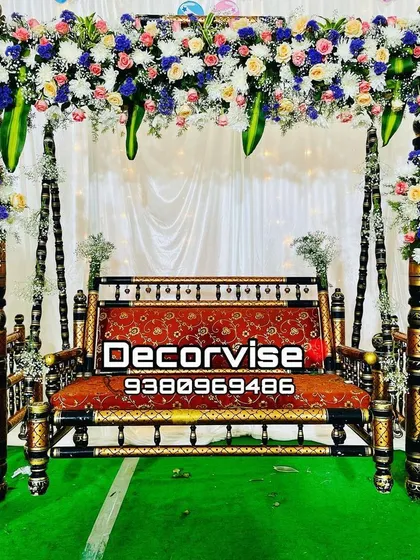A classic baby shower swing decoration featuring a mix of white, pink, peach, and blue flowers. The setup includes a simple white backdrop to highlight the colourful floral arrangements on the swing and side stands.
