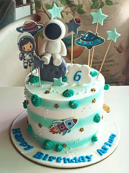 A charming astronaut-themed cake with striped buttercream, cute space explorers, a UFO, and a rocket ship, perfect for a 6th birthday.