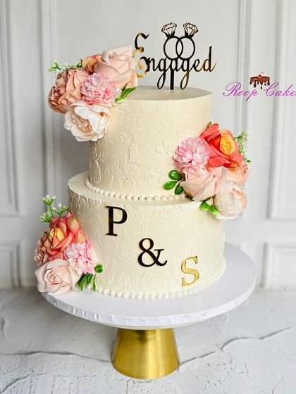 A masterpiece of love and elegance for an engagement. This two-tier cake is beautifully adorned with delicate flowers, a subtle stencil texture, and personalized with the couple's names.