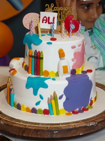 A close-up of the amazing two-tiered art-themed cake, decorated with edible crayons, paint splatters, and glue bottles.