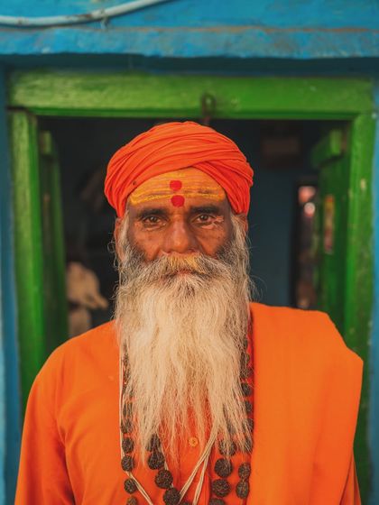 A close-up portrait of a sadhu in Varanasi. His gaze is direct, and his face, marked with tilak, tells a story of devotion and resilience against the vibrant blue and green of a local doorway.