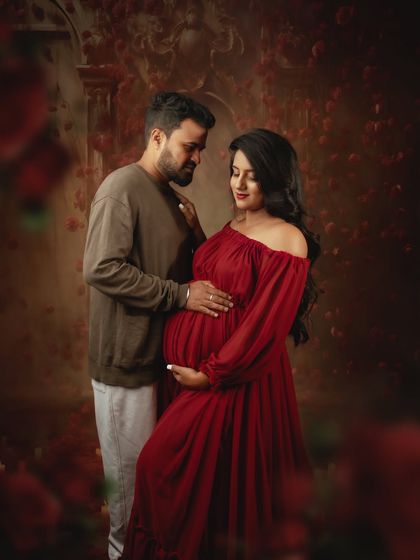 The partner's gentle touch on her shoulder adds a layer of connection to this moody studio portrait. The deep red dress stands out beautifully against the dark, floral background.