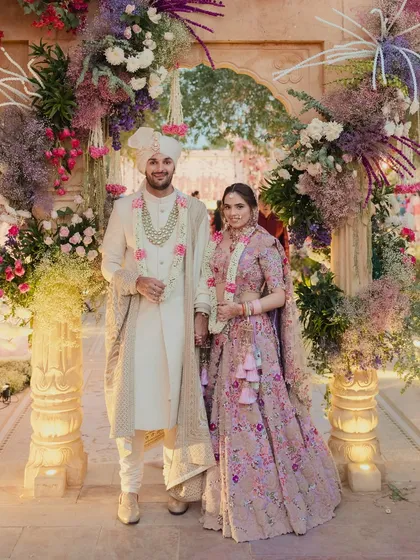 The couple stands under a lush floral arch, perfectly framed. The soft, multi-hued pastel flowers create a romantic and picturesque moment.