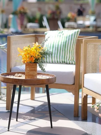 A cozy poolside lounge nook with light wood and cane chairs, a striped green cushion, and tropical floral accents.